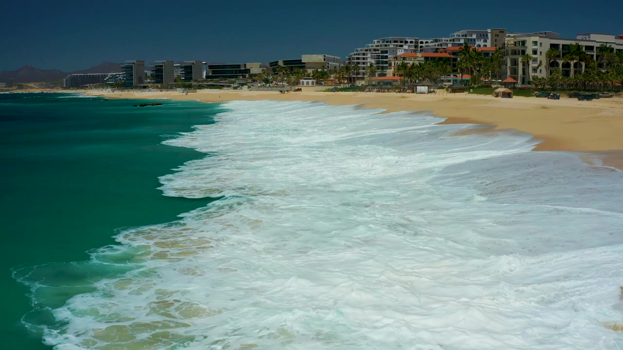 Midway through the clip, the foam line curves along the Dreams Los Cabos shoreline, with turquoise water on the left, wet sand on the right, and resort buildings behind the palms, with no visible people.