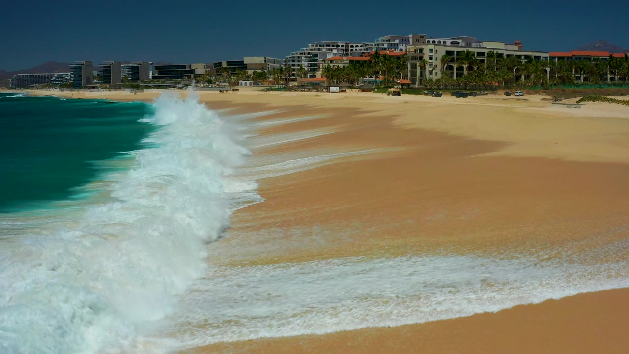 At Dreams Los Cabos in Baja California Sur, white waves push up a tan sandy beach with turquoise water at left, resort buildings and palms across the background, and no visible people.
