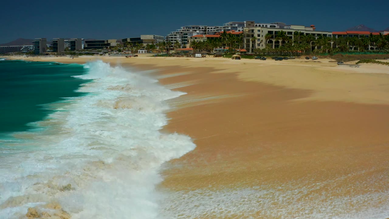 White surf spreads wider across the tan sand at Dreams Los Cabos, with clear turquoise water at left, beachfront resort structures along the upper edge, and no visible people.