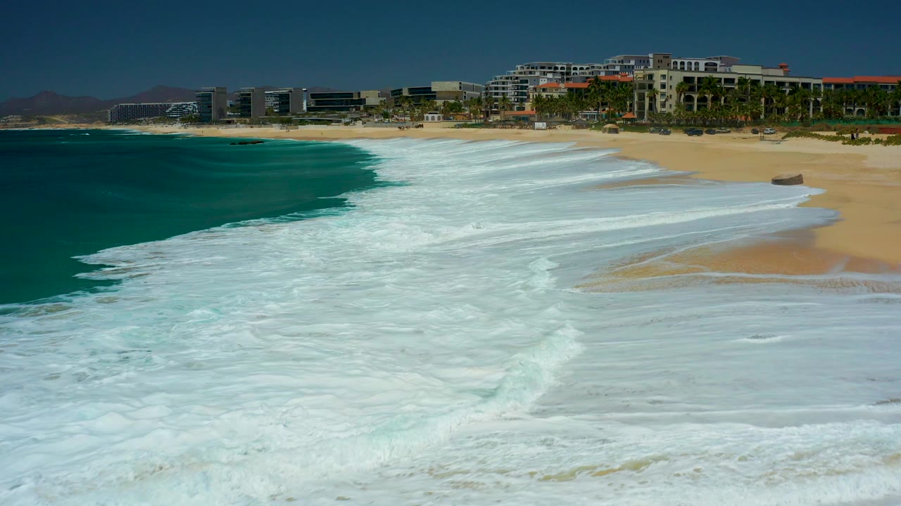 At Dreams Los Cabos in Baja California Sur, white surf covers much of the foreground beach while turquoise water, palm trees, resort buildings, distant hills, and no visible people fill the frame.