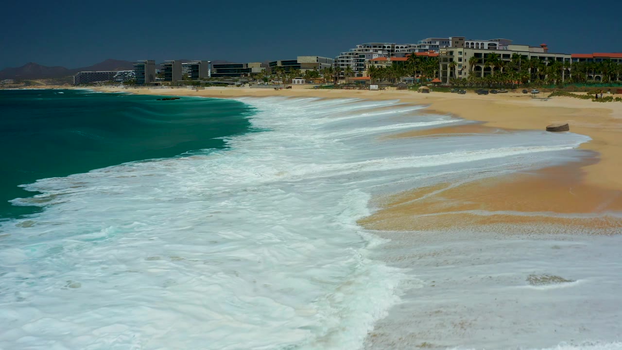 At Dreams Los Cabos in Baja California Sur, broken white surf reaches the beach in layered bands, with dry sand to the right, teal water to the left, resort buildings in back, and no visible people.