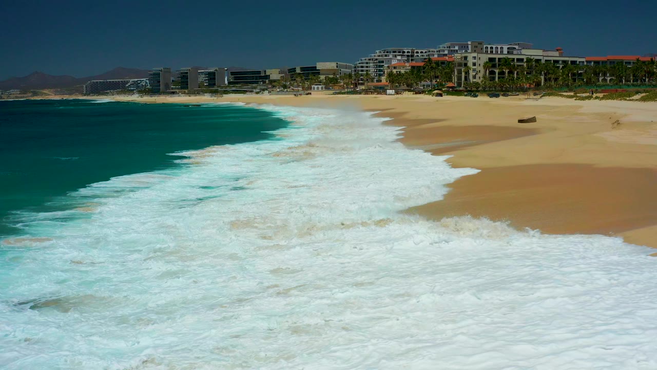 At Dreams Los Cabos in Baja California Sur, white surf spreads across the lower frame while turquoise water meets golden sand and resort buildings sit in the distance, with no visible people.