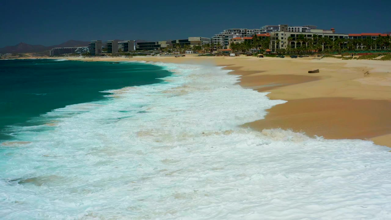 At 0:01, the white wave foam pushes higher across the sand at Dreams Los Cabos, leaving a broad surf band between the turquoise sea and the resort buildings, with no visible people.