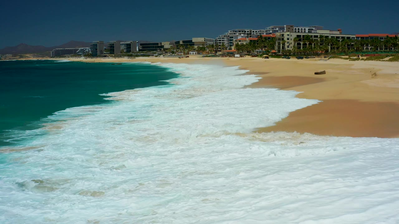 At 0:02, a larger sheet of whitewater covers most of the foreground beach at Dreams Los Cabos while the exposed sand narrows on the right, with no visible people.