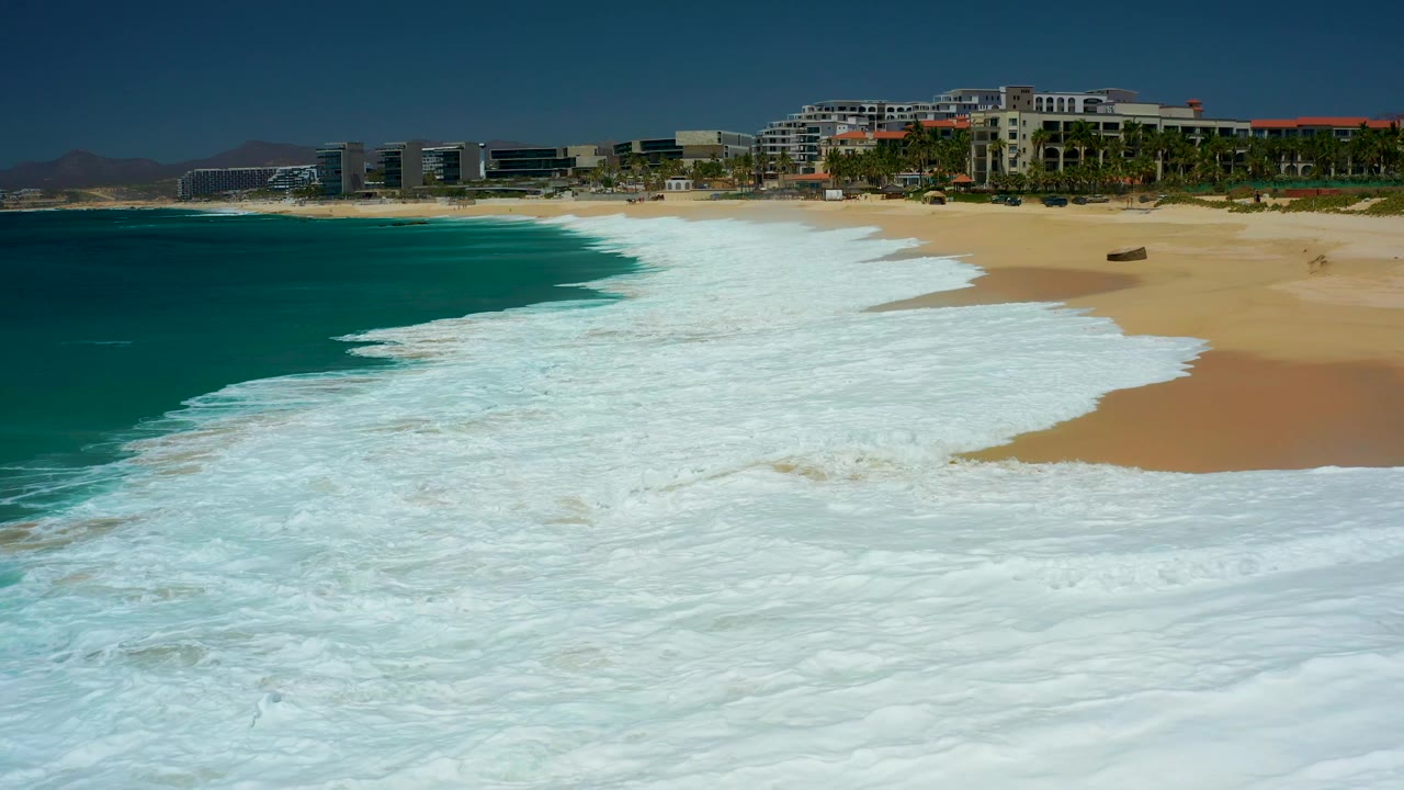 Ocean foam recedes across the beach with buildings and palms beyond the sand.