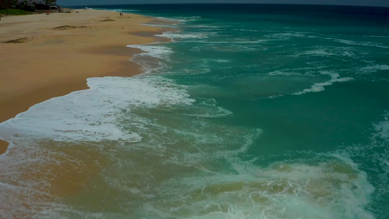 A broad sheet of white foam advances farther up the sandy shoreline at Dreams Los Cabos, with open turquoise water filling the right side and no visible people.