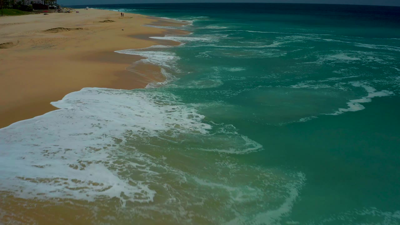 The surf forms a curved white edge along the golden beach in Dreams Los Cabos, while darker turquoise water sits beyond the breaking foam and no visible people appear.