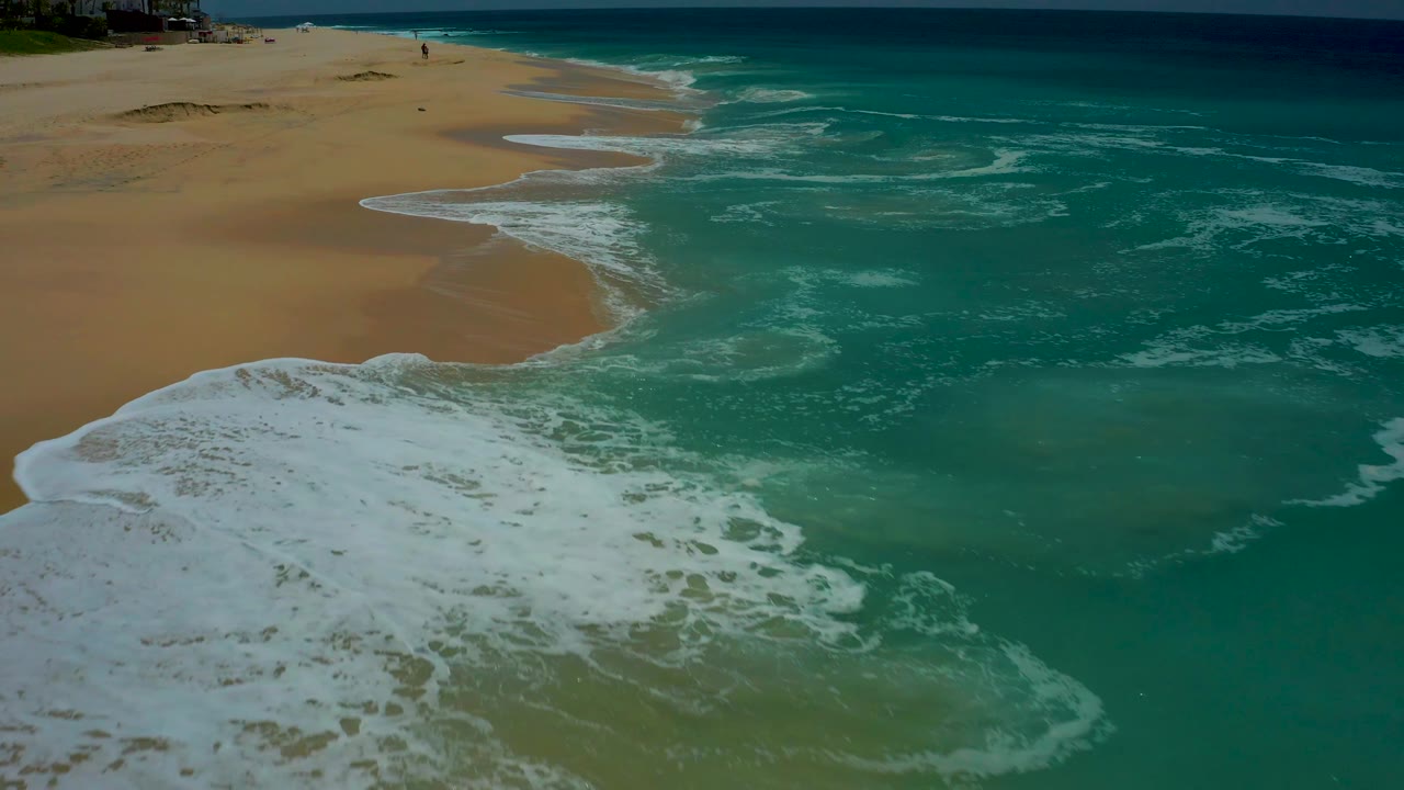 White foam trails pull back from the sand at Dreams Los Cabos, leaving darker wet areas near the waterline and clear turquoise surf across the frame.