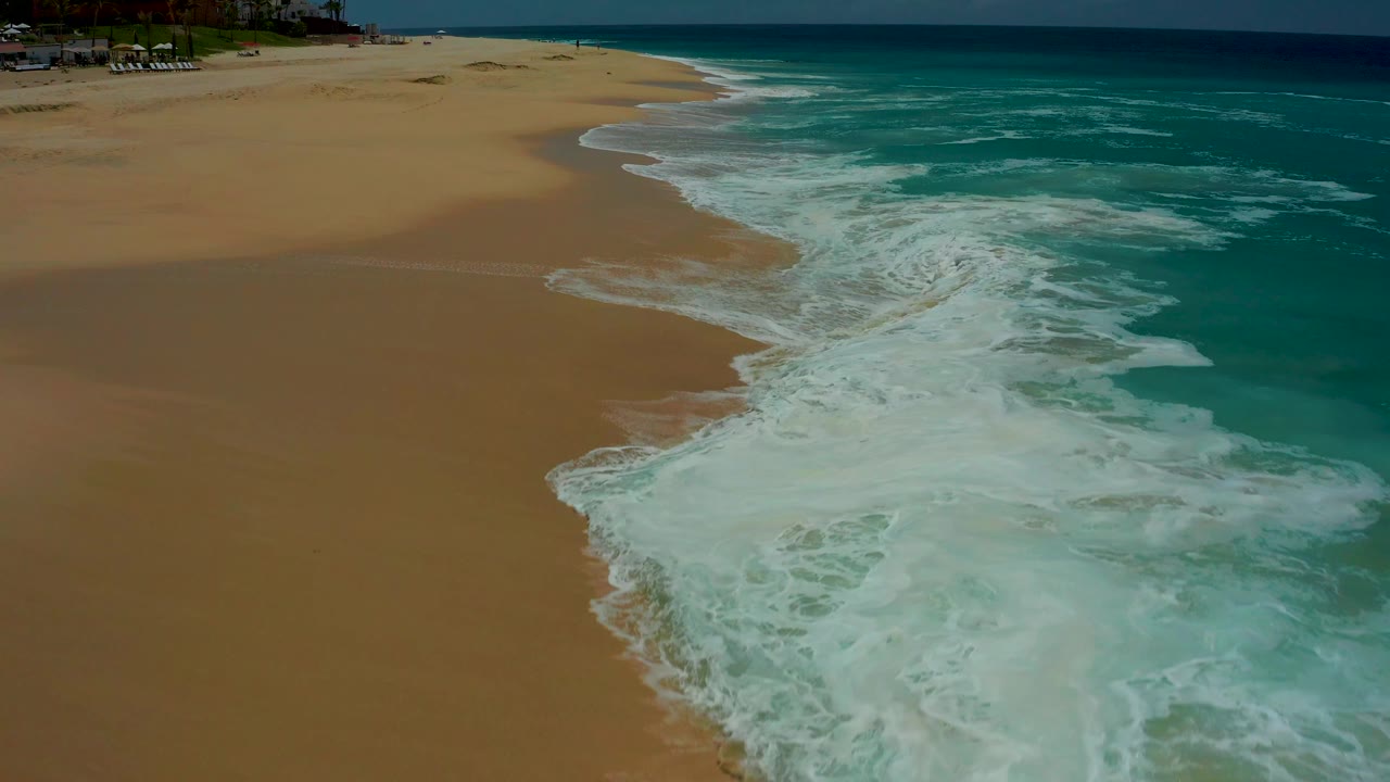 At 0:01, the Dreams Los Cabos shoreline shows a broader band of whitewater pushing across wet golden sand, with open turquoise water and no visible people.