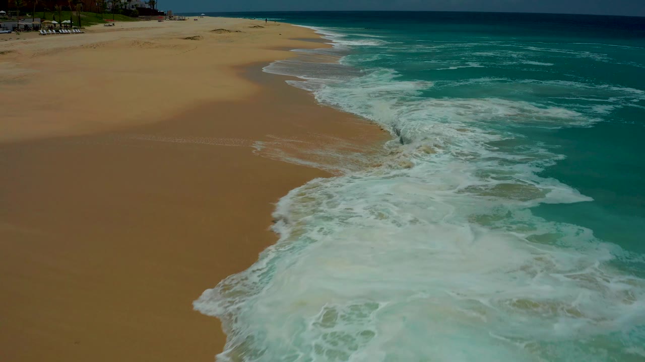 At 0:02, a curved break of white foam marks the edge of the turquoise ocean against the sandy Dreams Los Cabos beach, with no visible people in the frame.