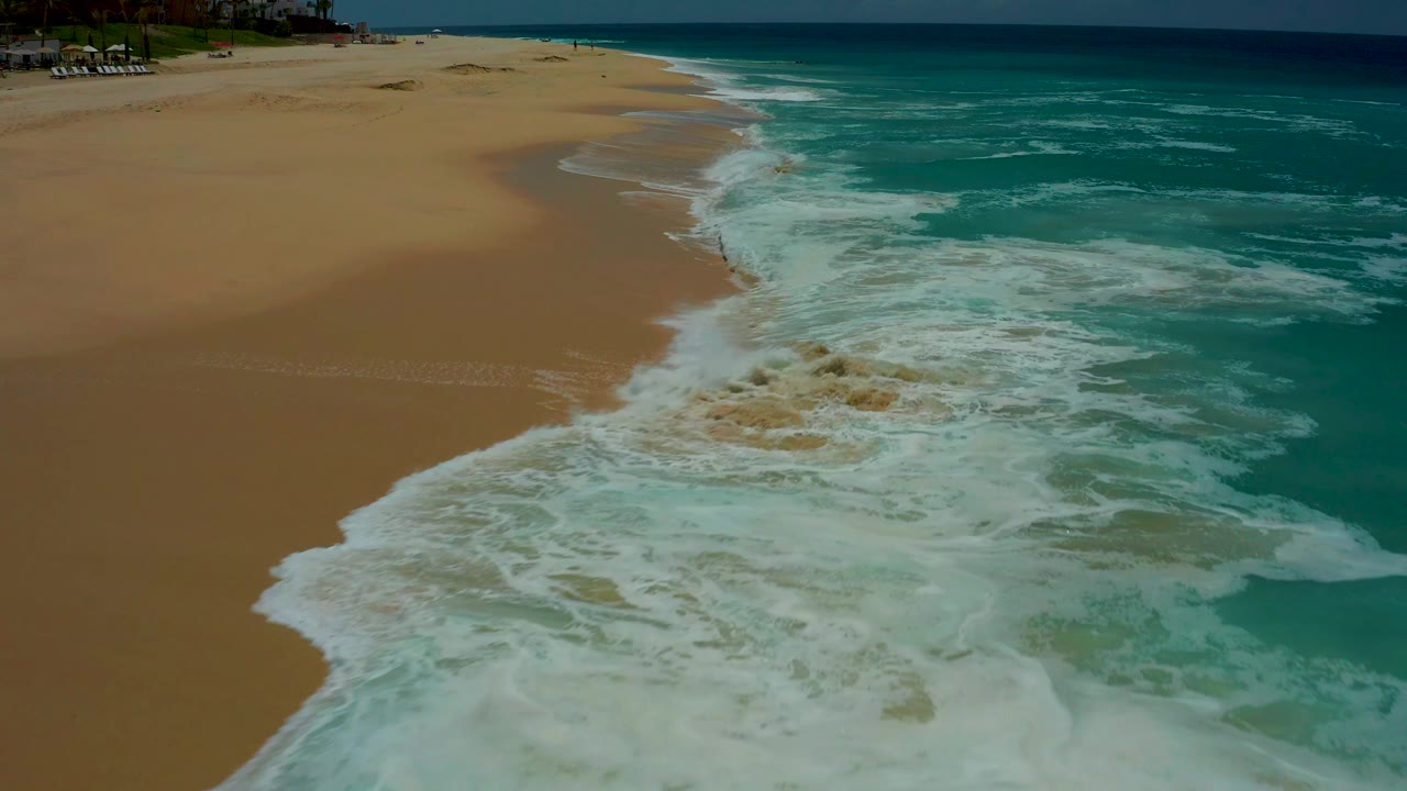 At 0:03, the aerial view looks along the Dreams Los Cabos beach as white surf rolls up over tan sand and turquoise water fills the right side, with no visible people.