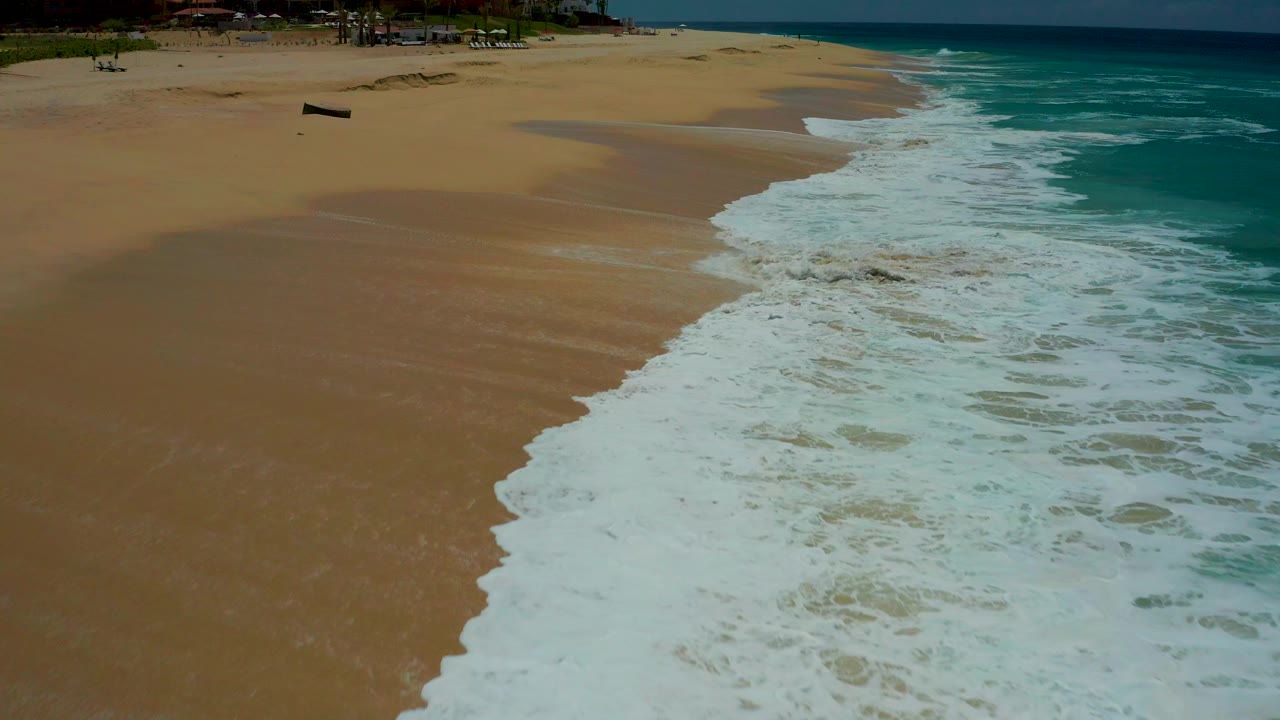 Aerial view of white surf reaching a sandy beach beside turquoise water.