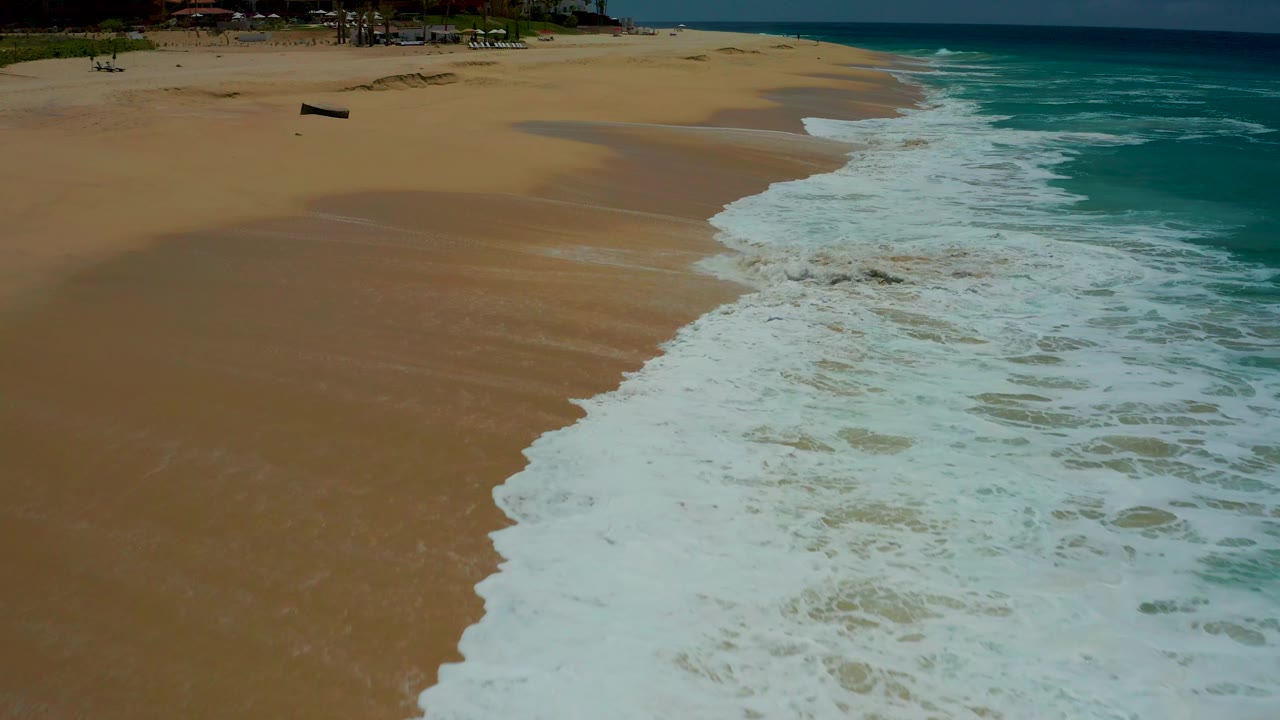 An overhead frame at Dreams Los Cabos shows a broad white wave wash pushing farther up the wet sand, with clear turquoise water to the right and no visible people.