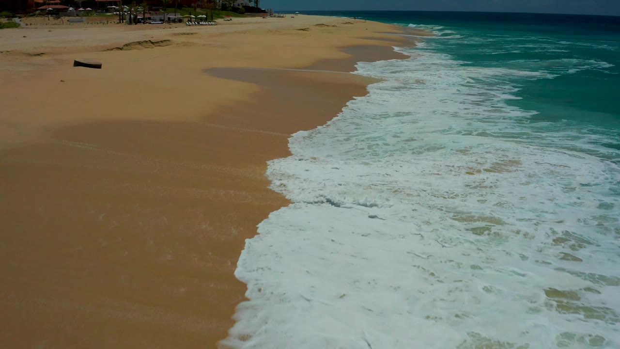 The aerial view shows the wave edge forming a diagonal white line across the Dreams Los Cabos sand, with darker wet patches near the upper beach and no visible people.