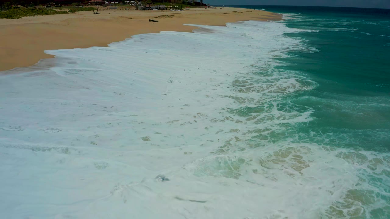 White surf spreading across a sandy beach beside turquoise ocean.