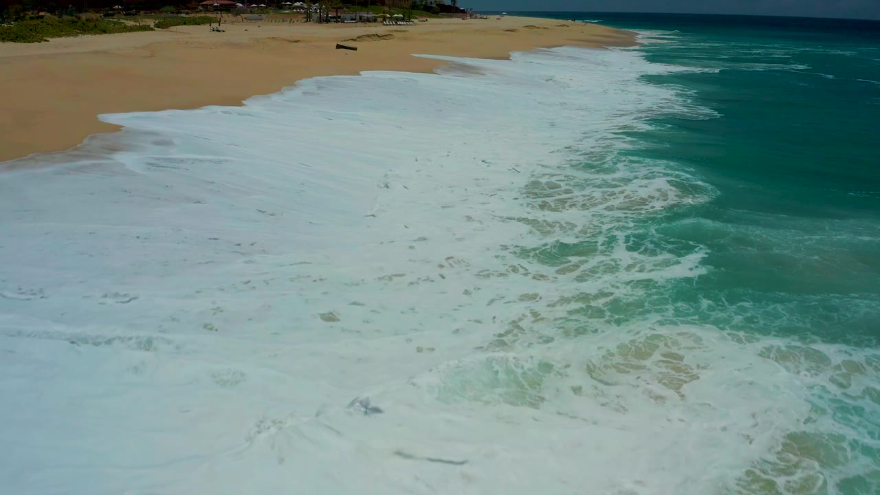 The aerial view holds over the Dreams Los Cabos shoreline as white wash runs diagonally across wet tan sand with turquoise ocean at right and no visible people.