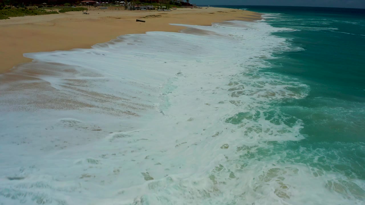 Foam begins to thin across the Dreams Los Cabos beach, revealing more wet sand while the darker turquoise sea remains along the right edge and no visible people are present.