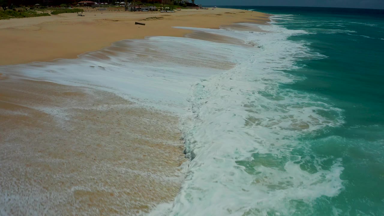Aerial shoreline detail at Dreams Los Cabos shows the foam line narrowing as wet sand opens across the left half of the frame, with no visible people.
