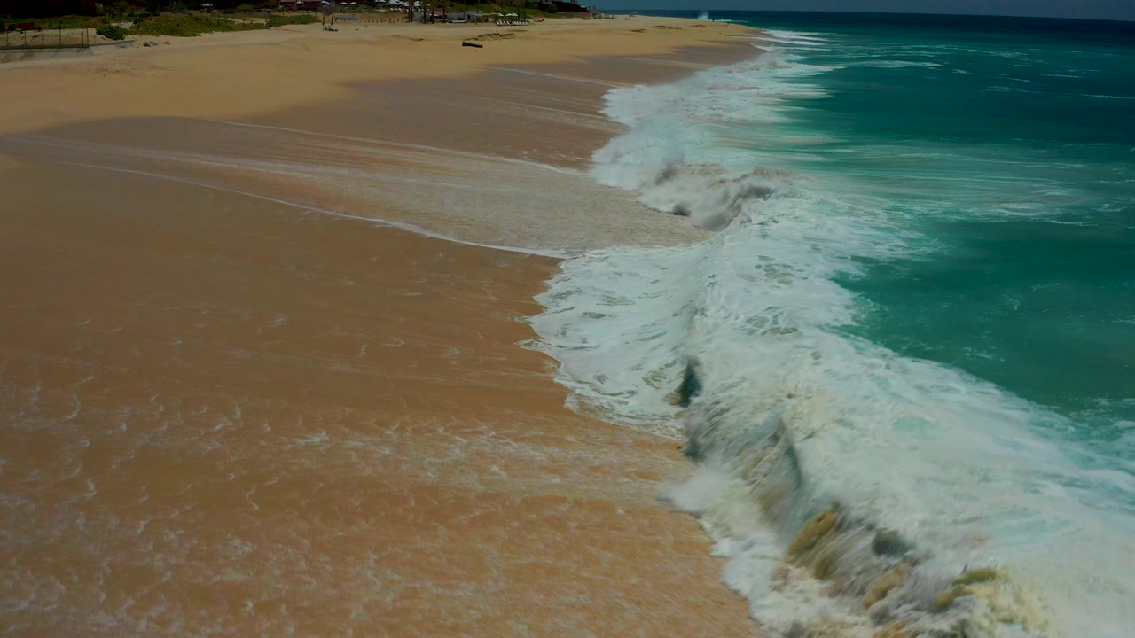 Aerial view at Dreams Los Cabos showing turquoise water breaking into white foam along a sandy beach, with no visible people.