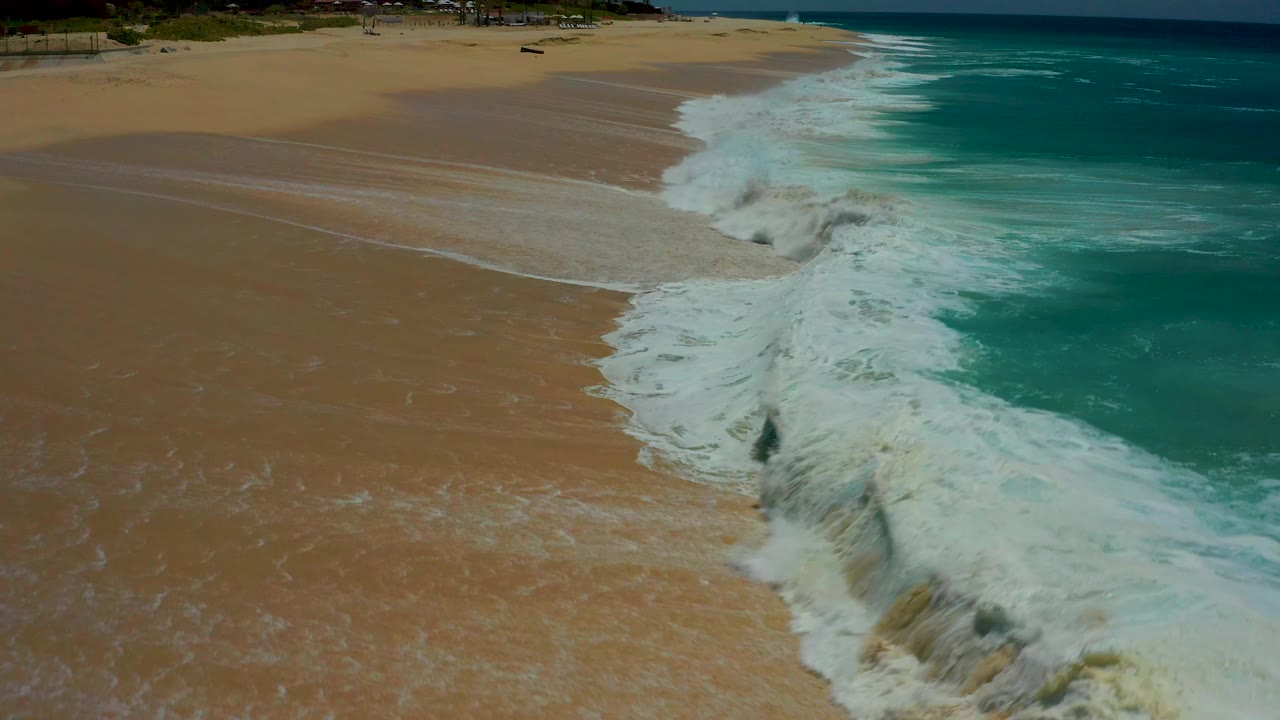 At Dreams Los Cabos, white surf curls diagonally onto wet sand while turquoise water holds the right side of the frame, with no visible people.