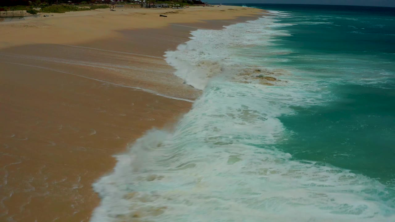 Foamy whitewater advances across the sandy Dreams Los Cabos shoreline as deeper turquoise water remains offshore, with no visible people.