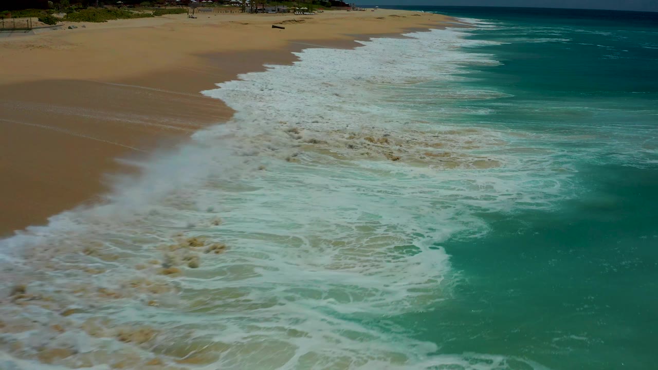 A broad sheet of white surf covers much of the lower Dreams Los Cabos beach while exposed sand remains toward the upper left, with no visible people.