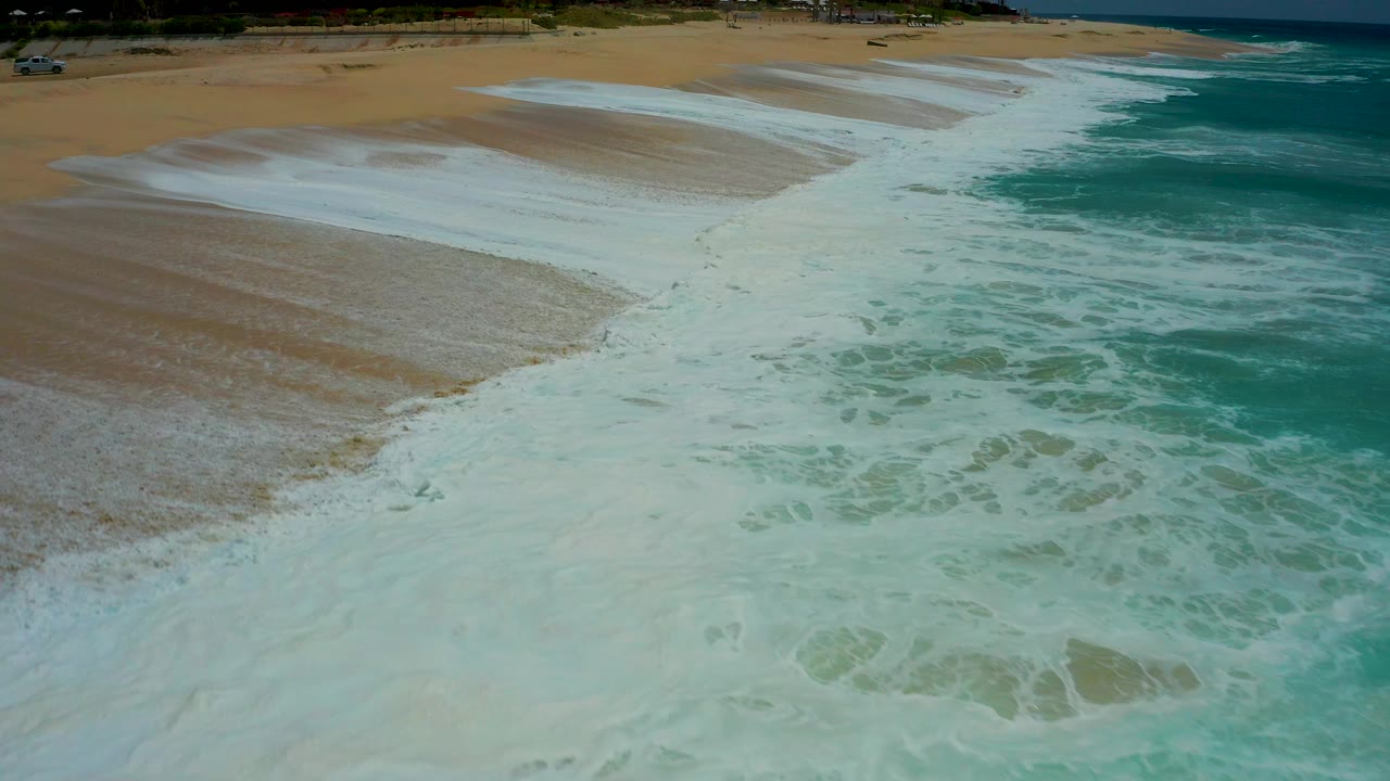 At Dreams Los Cabos in Baja California Sur, white foam covers the lower beach as turquoise water sits to the right and no visible people appear in frame.