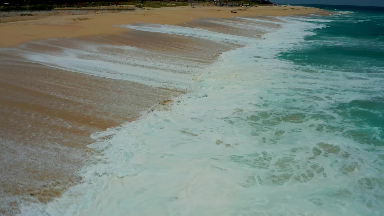 The wave line curves across the sand while white foam breaks into the turquoise water at Dreams Los Cabos, with the upper beach faintly visible and no visible people.