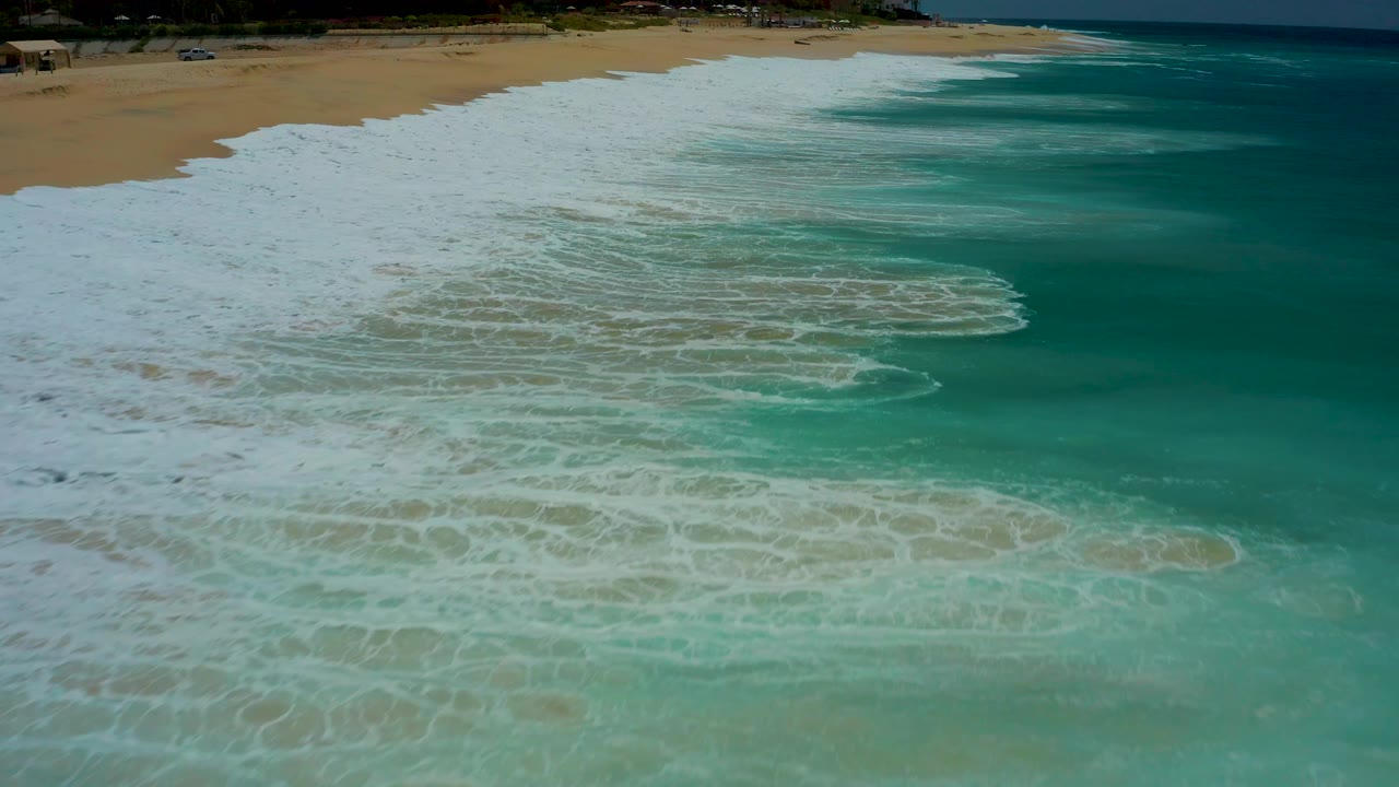 Aerial surf frame at Dreams Los Cabos with white foam covering more of the shallow turquoise water and the sandy beach running across the top, no visible people.