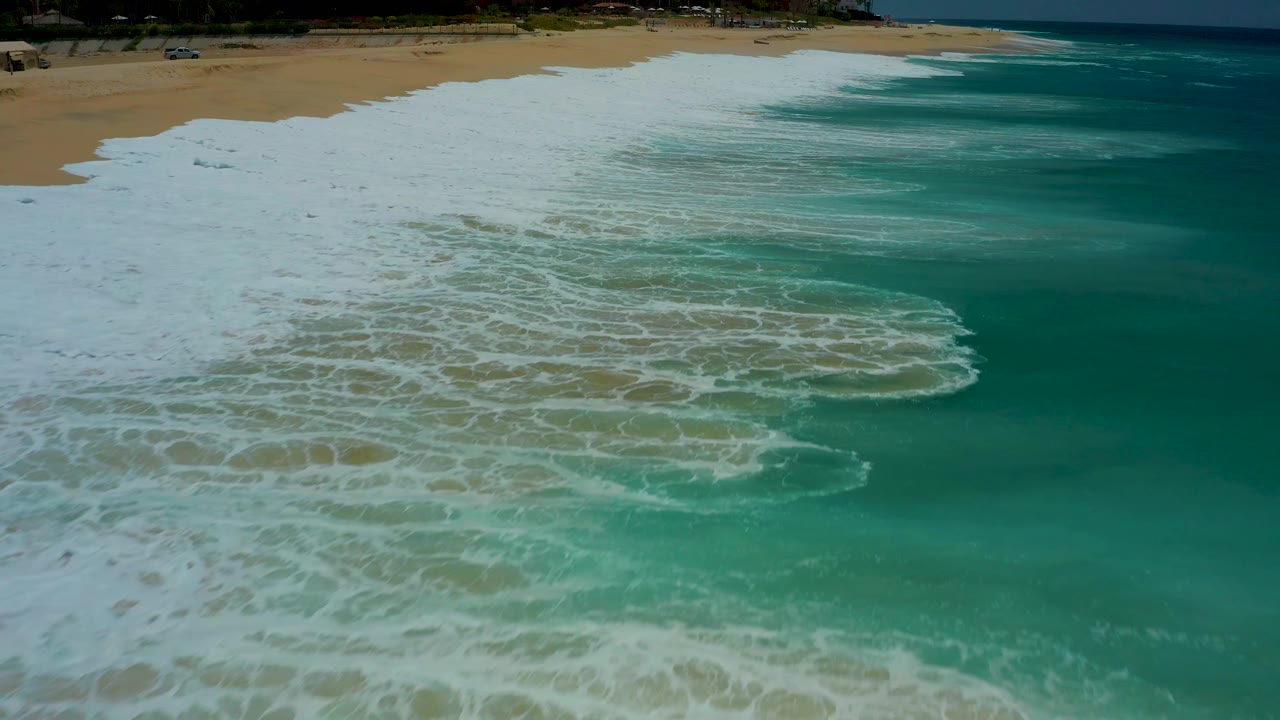 Aerial view at Dreams Los Cabos with a curved band of white breaking foam across green-blue water and a sandy beach strip in the background, no visible people.