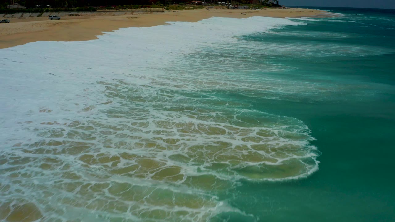Aerial frame at Dreams Los Cabos showing dense white foam filling the lower left and center while turquoise water remains on the right, with no visible people.