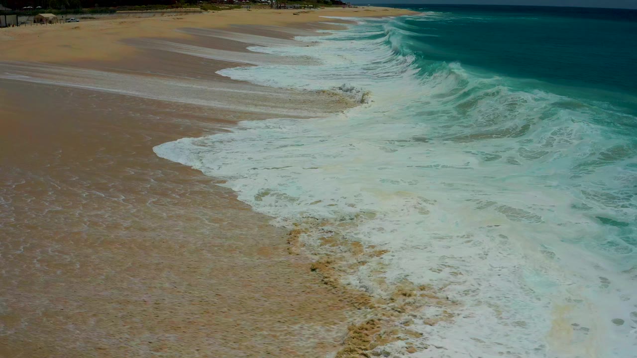 Aerial shoreline frame at Dreams Los Cabos showing foamy water widening over the sand while darker blue ocean sits beyond, no visible people.