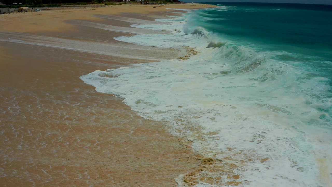 Aerial view at Dreams Los Cabos where a curling breaker meets the shoreline and leaves streaks of white foam over tan wet sand, no visible people.