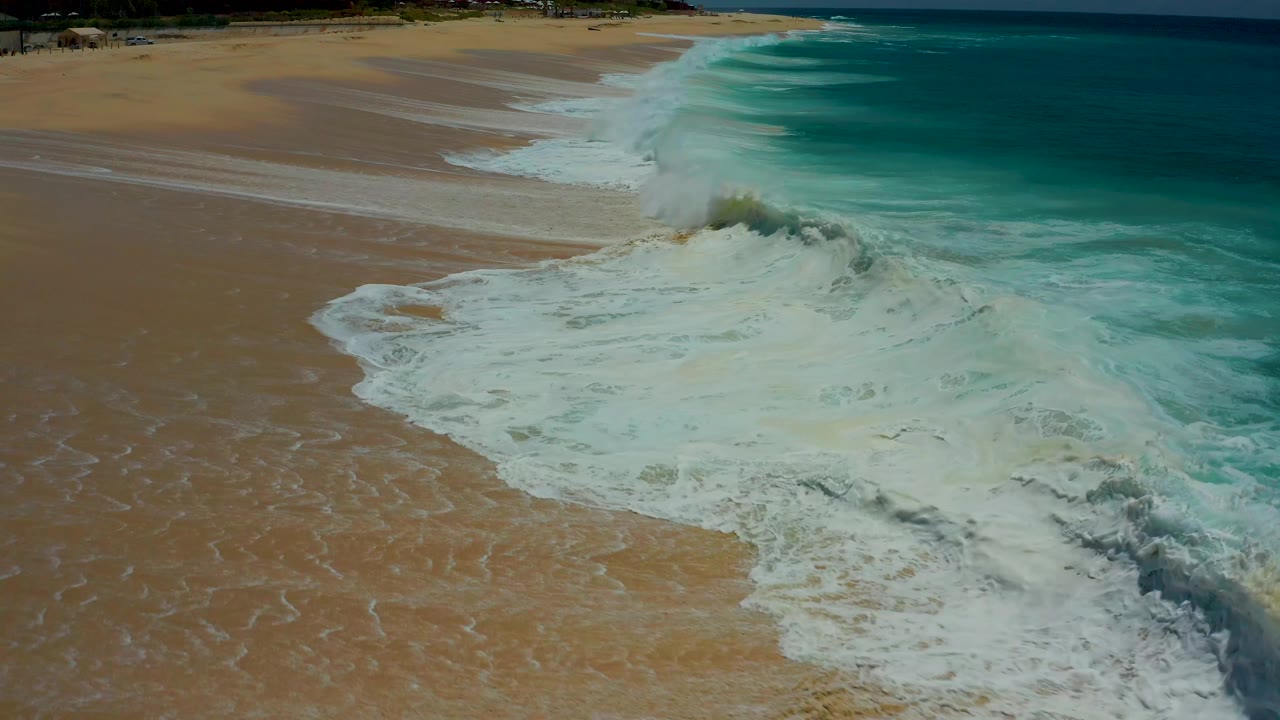 Aerial Dreams Los Cabos surf frame with a compact breaker near the center and white foam spreading across the beach side of the shoreline, no visible people.
