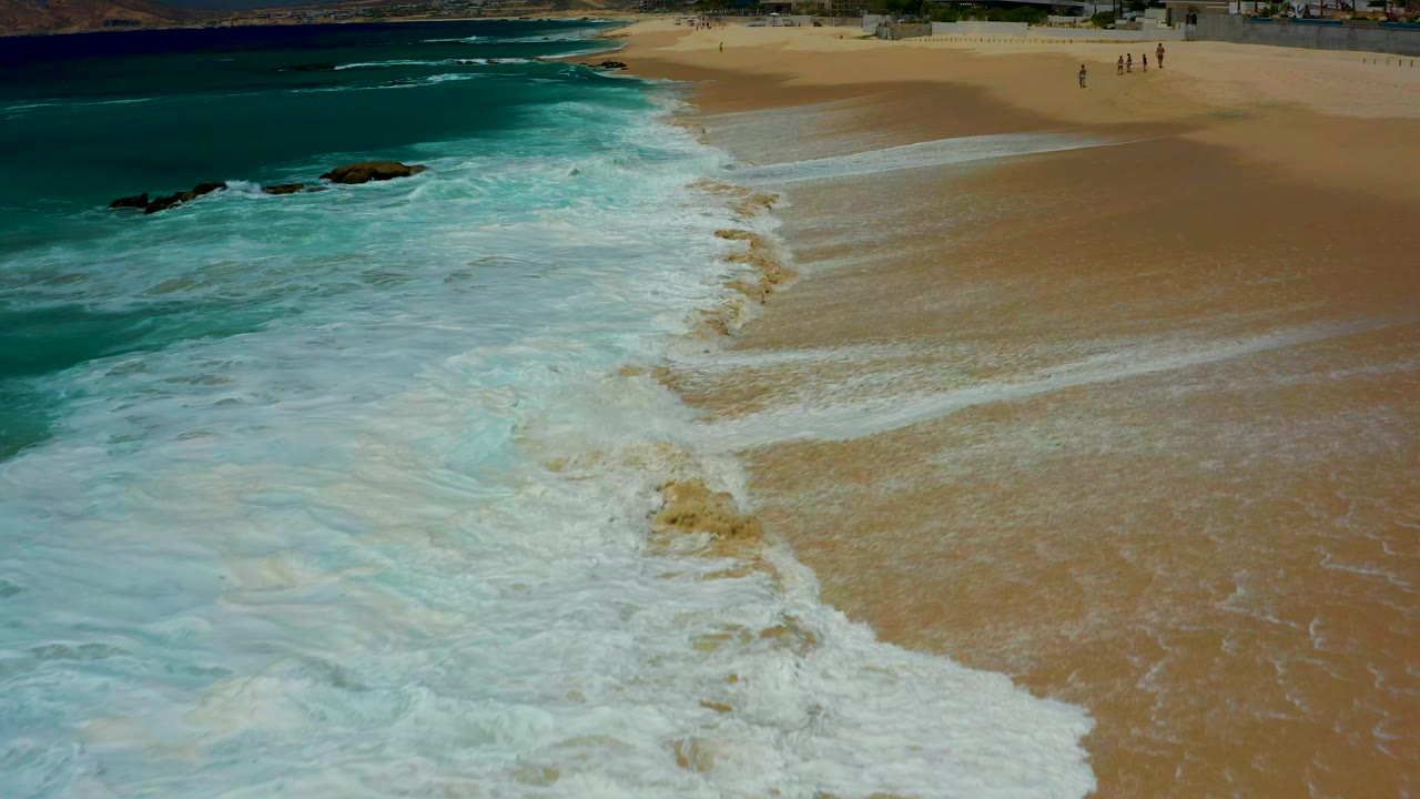 Aerial frame from Dreams Los Cabos showing turquoise surf spreading white foam across tan beach sand, with small dark rocks near the left side and distant people near the upper right.