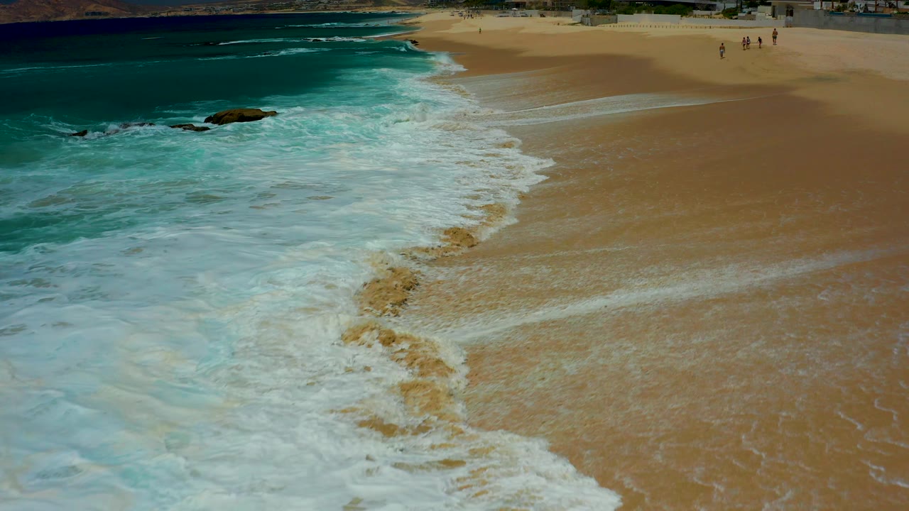 Aerial frame from Dreams Los Cabos where white foam forms a broad curved edge across wet sand, with turquoise sea to the left and distant people grouped high on the beach.