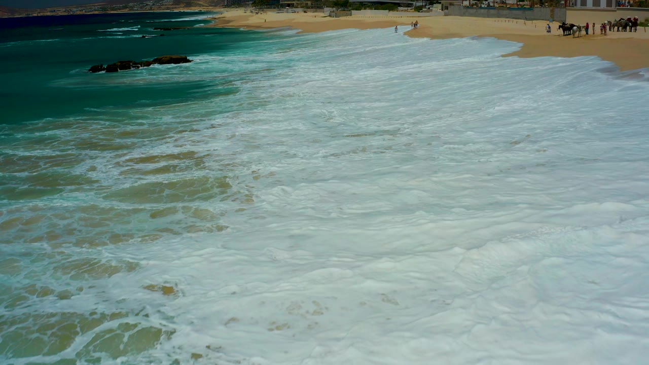 At Dreams Los Cabos in Baja California Sur, Mexico, foamy surf covers most of the foreground sand while dark rocks sit offshore at upper left and small distant people appear near the upper beach.