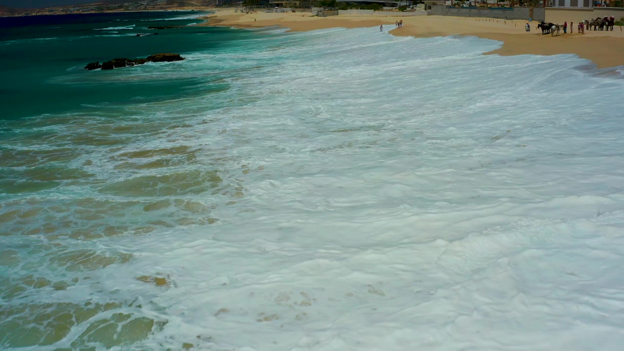 White surf washes over tan sand near dark offshore rocks.