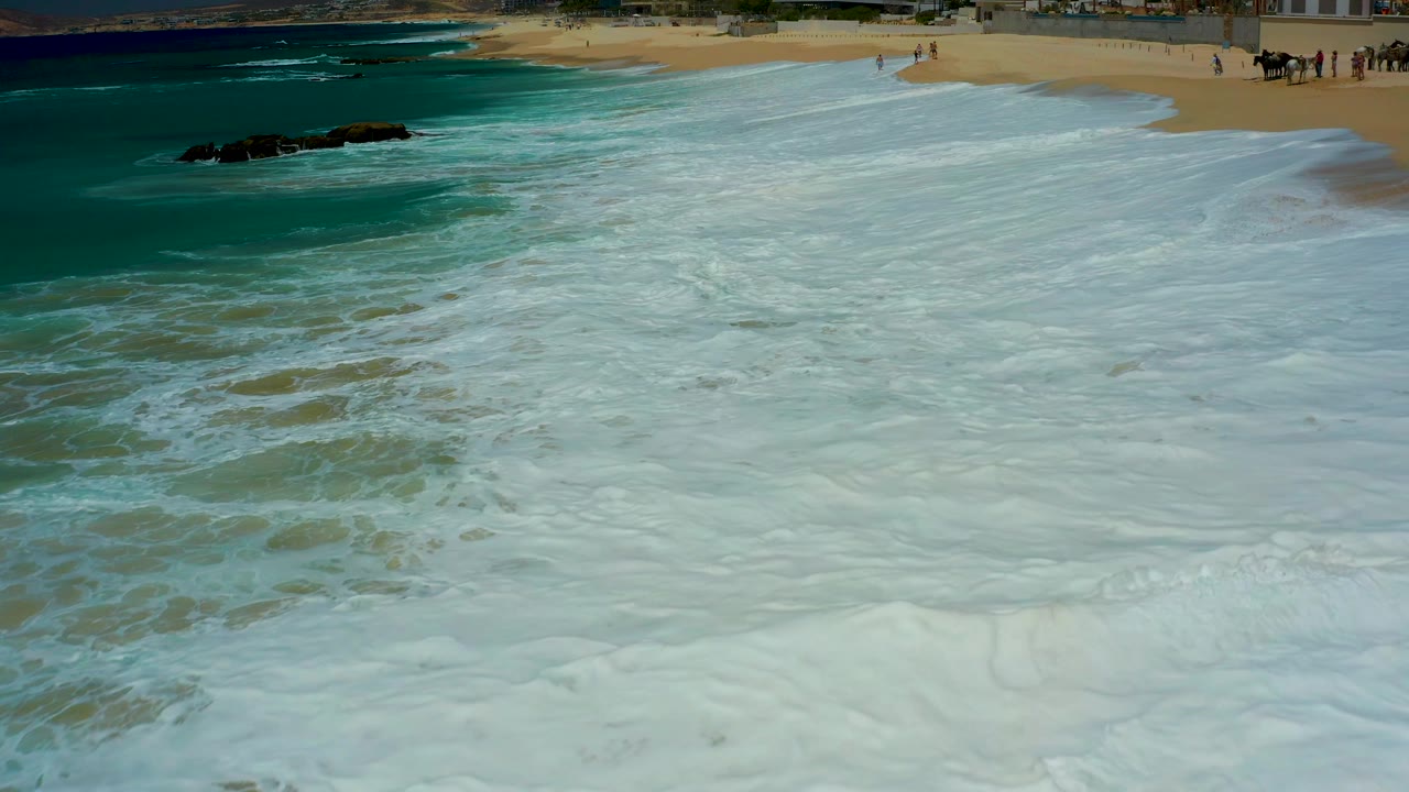 At Dreams Los Cabos in Baja California Sur, Mexico, the foamy water blankets the lower frame while a sandy strip and several small distant people remain visible near the top edge.