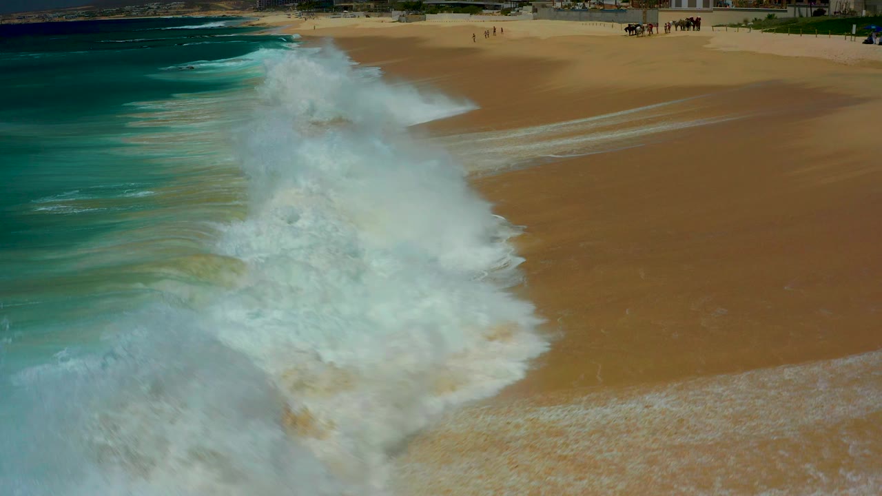 Aerial view at Dreams Los Cabos as a tall white wave breaks diagonally across tan sand, with blue-green ocean on the left and tiny people far up the beach.