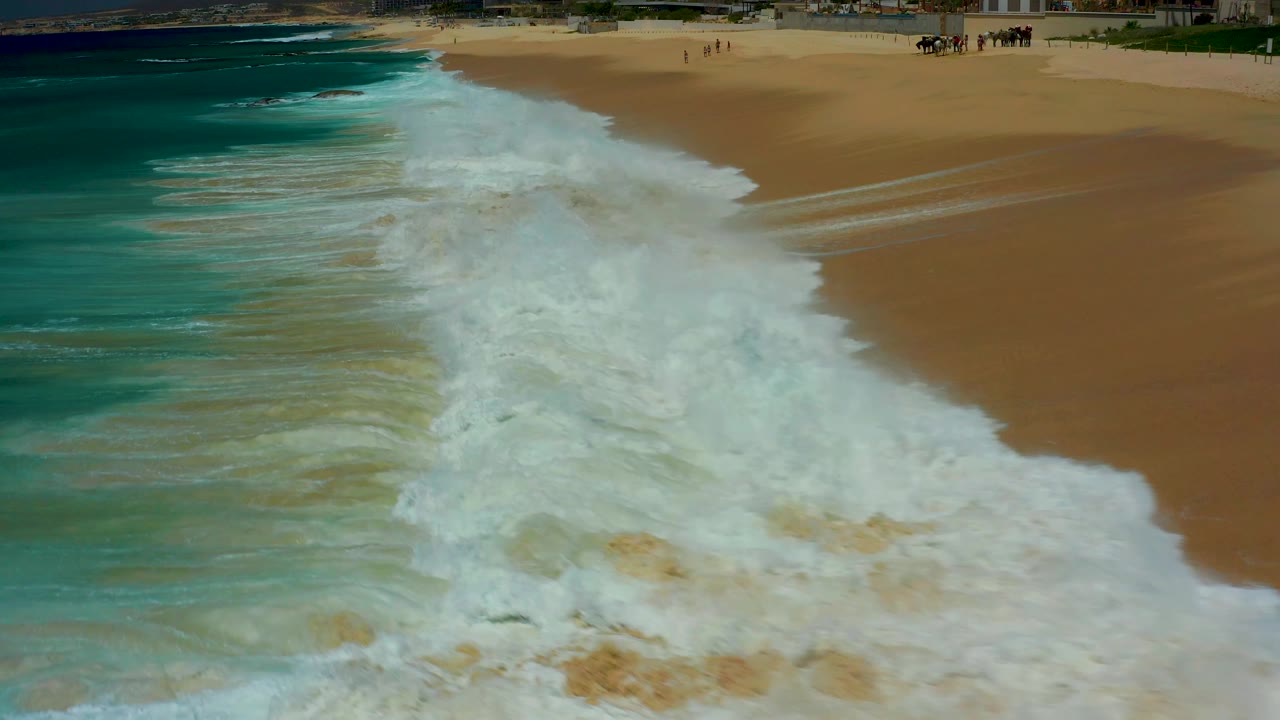 Aerial view at Dreams Los Cabos as white foam covers more of the lower frame, with tan beach still open at the top and small people visible in the distance.