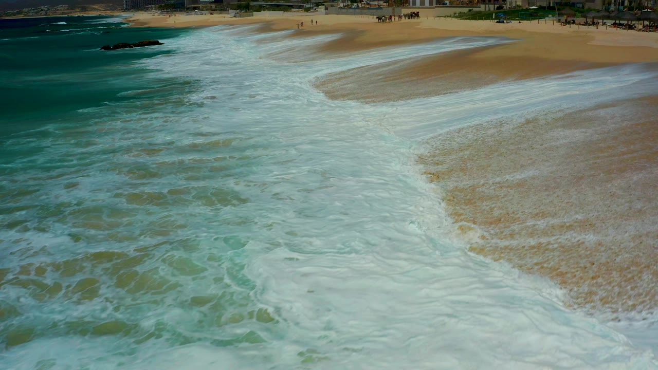 At Dreams Los Cabos in Baja California Sur, white foam covers much of the wet sand as turquoise water enters from the left, with no visible people in the surf area.
