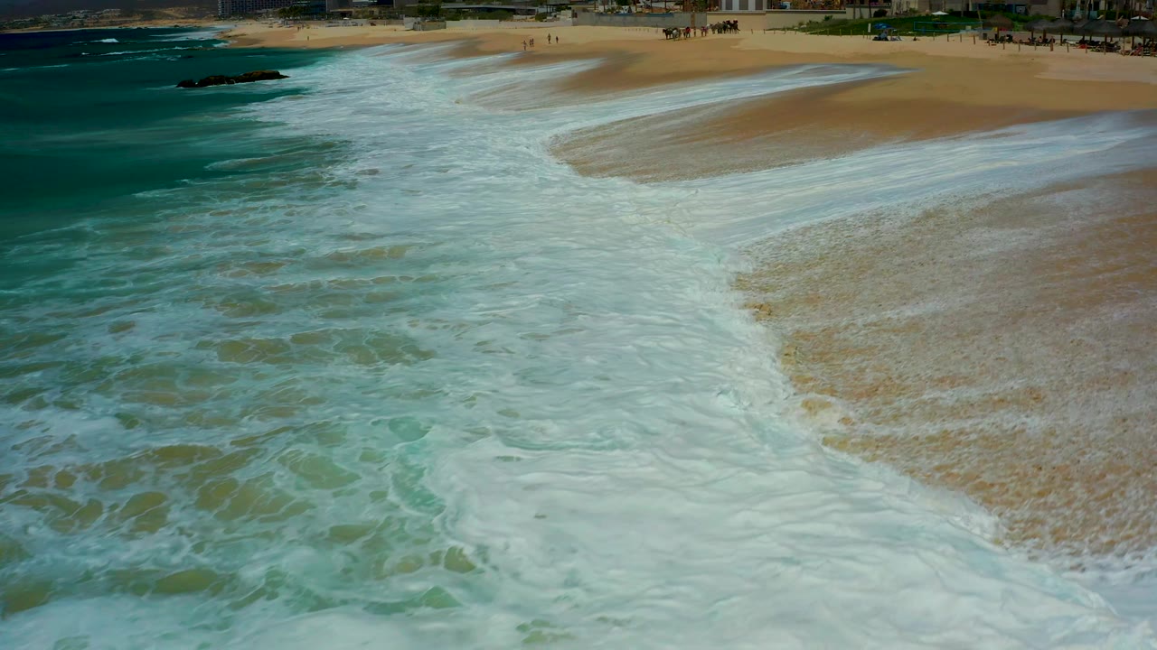 At 0:01, the foamy wash forms a broad diagonal band across the Dreams Los Cabos beach, with darker ocean water on the left and no visible people in the surf area.