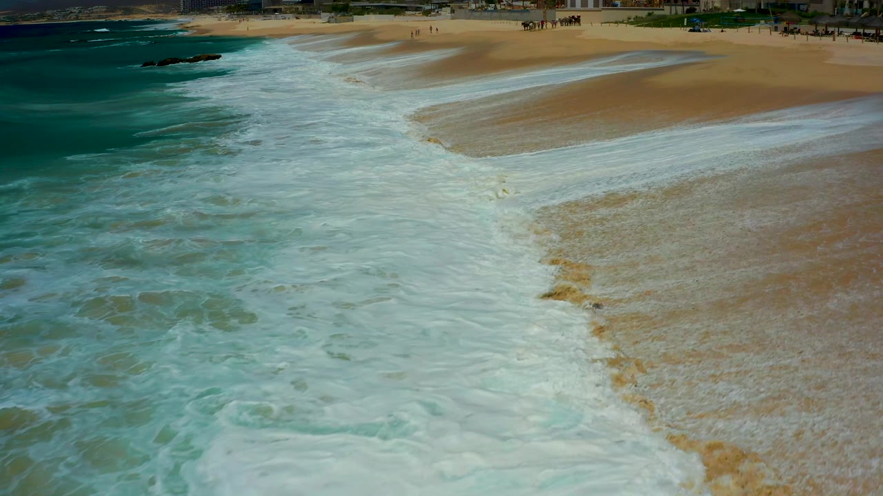 At Dreams Los Cabos, thinner white foam streaks across the sand while the darker teal ocean remains along the left edge, with no visible people in the surf area.