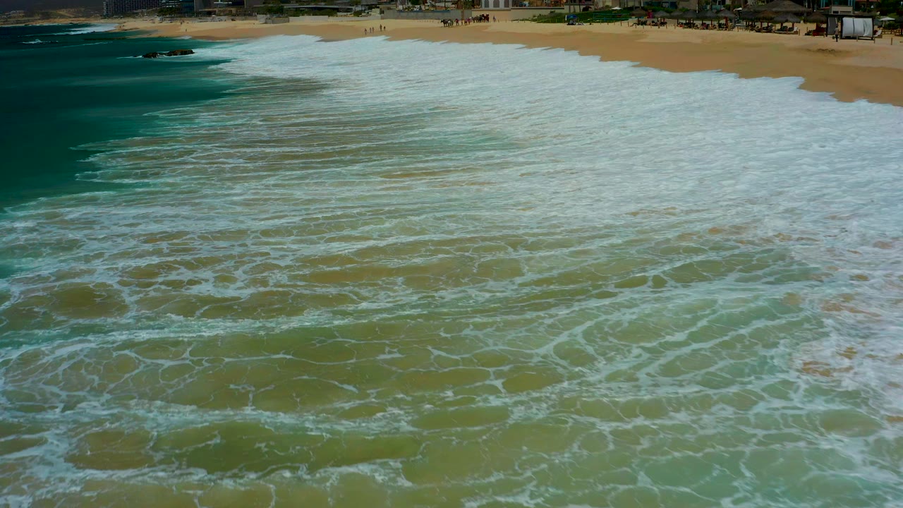 Overhead view at Dreams Los Cabos, Mexico, with teal ocean at left, white surf foam across green shallows, and a sandy beach along the top edge with no visible people.
