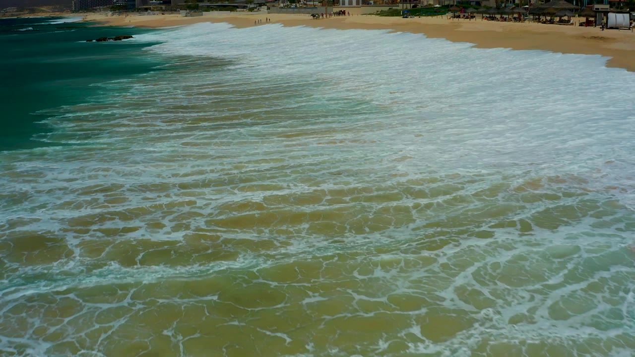 Overhead Dreams Los Cabos beach view with layered white foam bands over green water, darker teal ocean to the left, and a thin sandy shore at the top with no visible people.