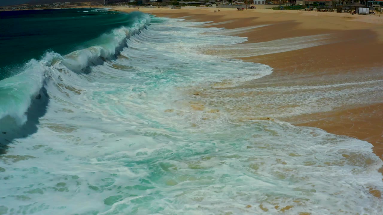 Aerial view at Dreams Los Cabos shows white foam widening across tan sand as a cresting wave runs diagonally along the shoreline, with no visible people in the frame.