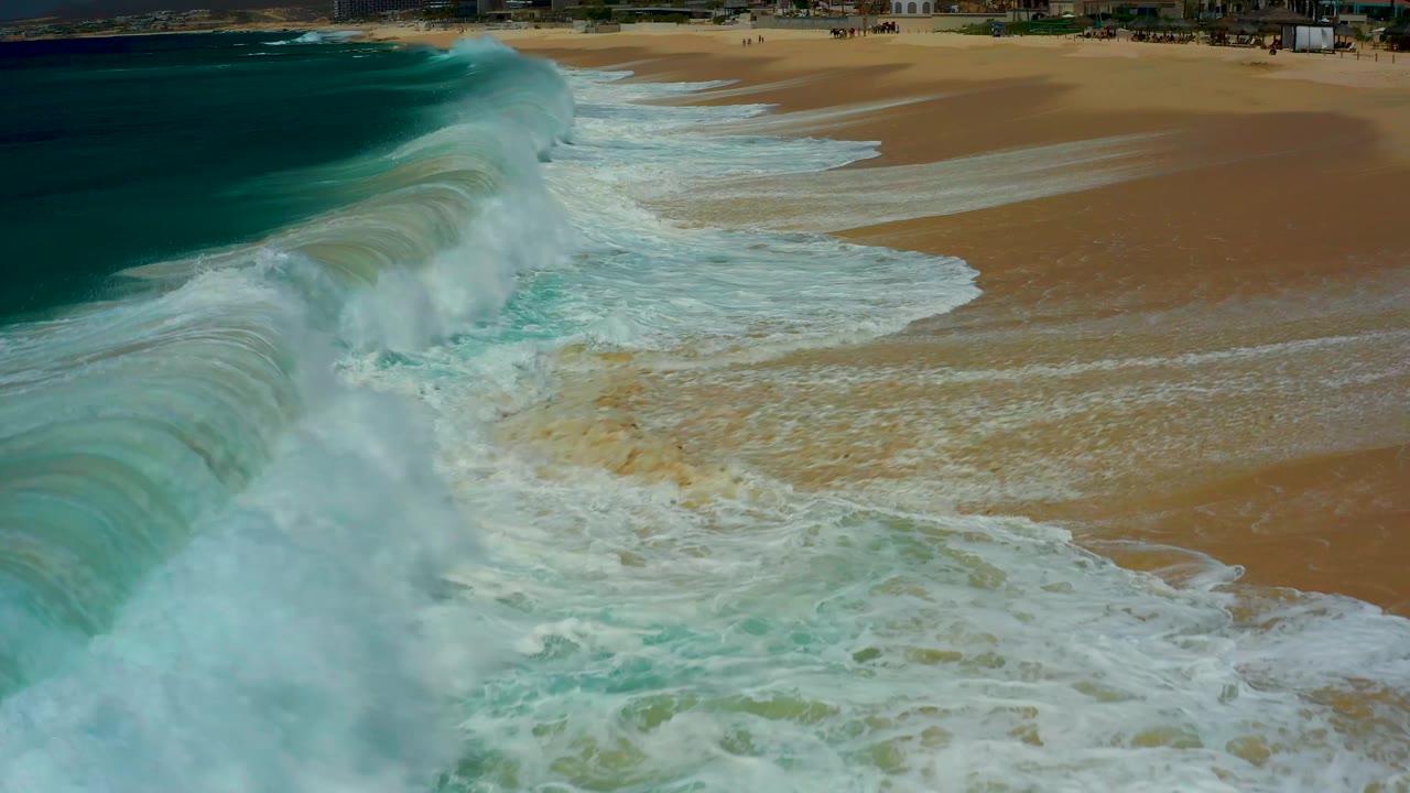 A curling turquoise wave rises near the left foreground at Dreams Los Cabos while sheets of white wash move across the sandy beach, with no visible people.