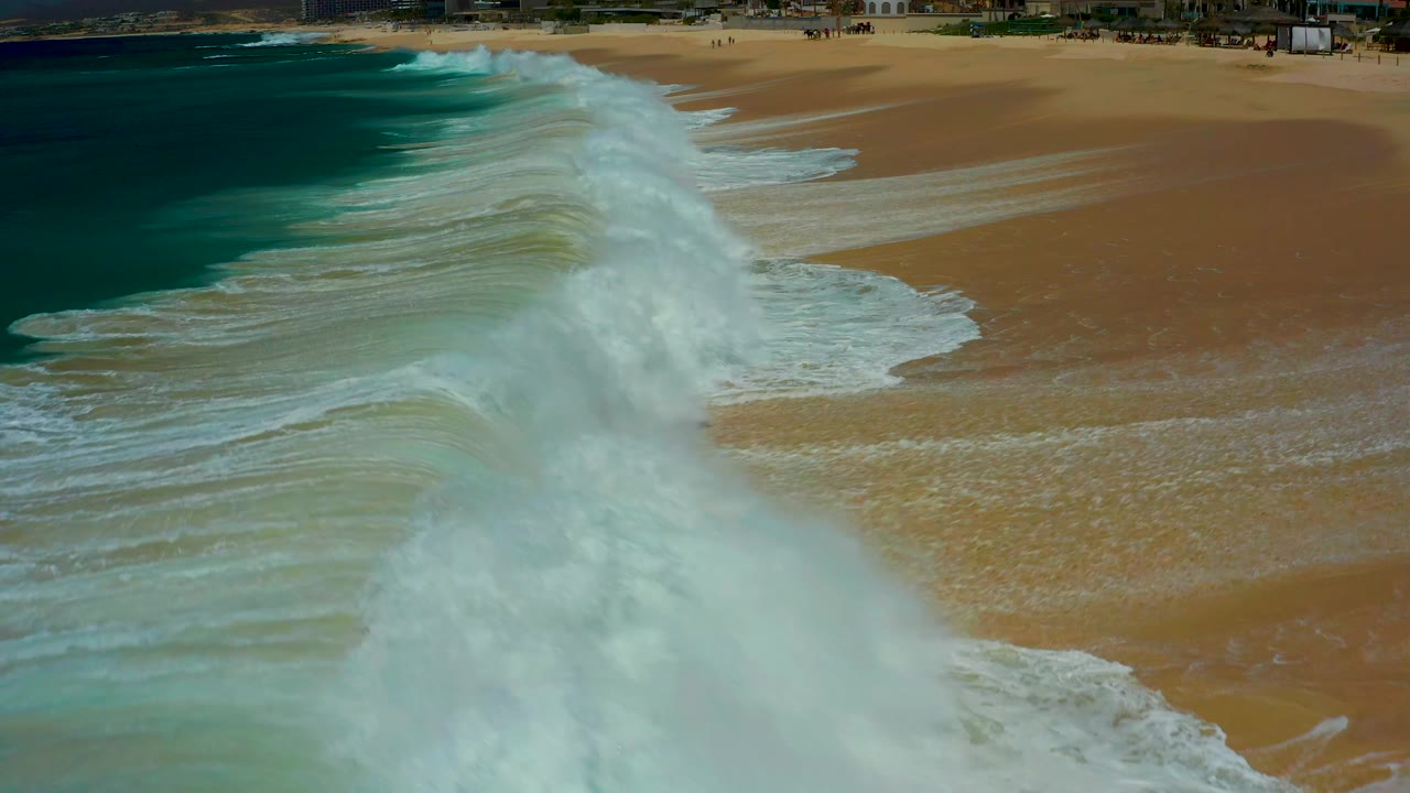 A broad plume of white surf moves across the sand at Dreams Los Cabos, with darker ocean water on the left and wet beach texture visible on the right, with no visible people.