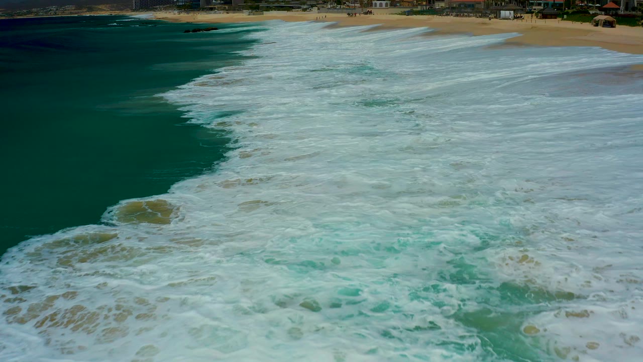 Aerial view at Dreams Los Cabos showing white surf spreading across teal water toward tan sand, with no visible people.